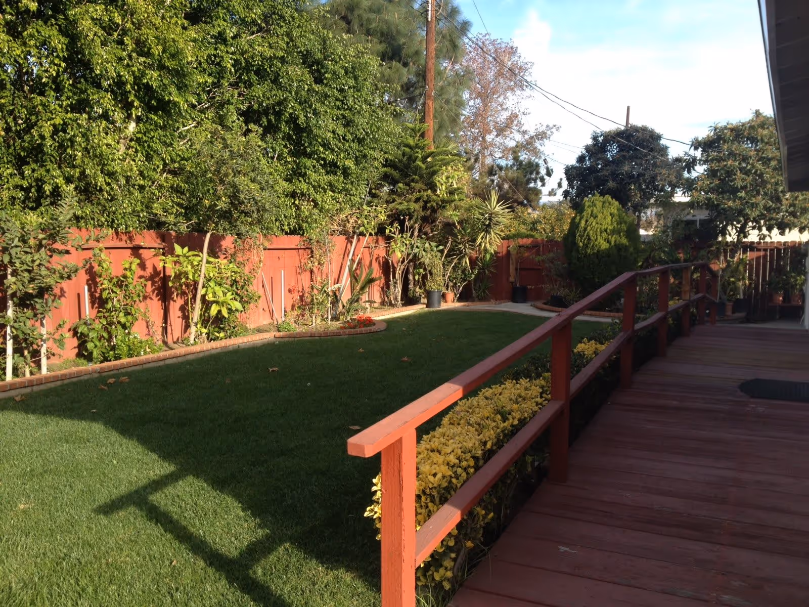 A well-maintained outdoor garden area with a green lawn, various plants and shrubs along a red wooden fence, and a wooden deck with a red railing on the right side.