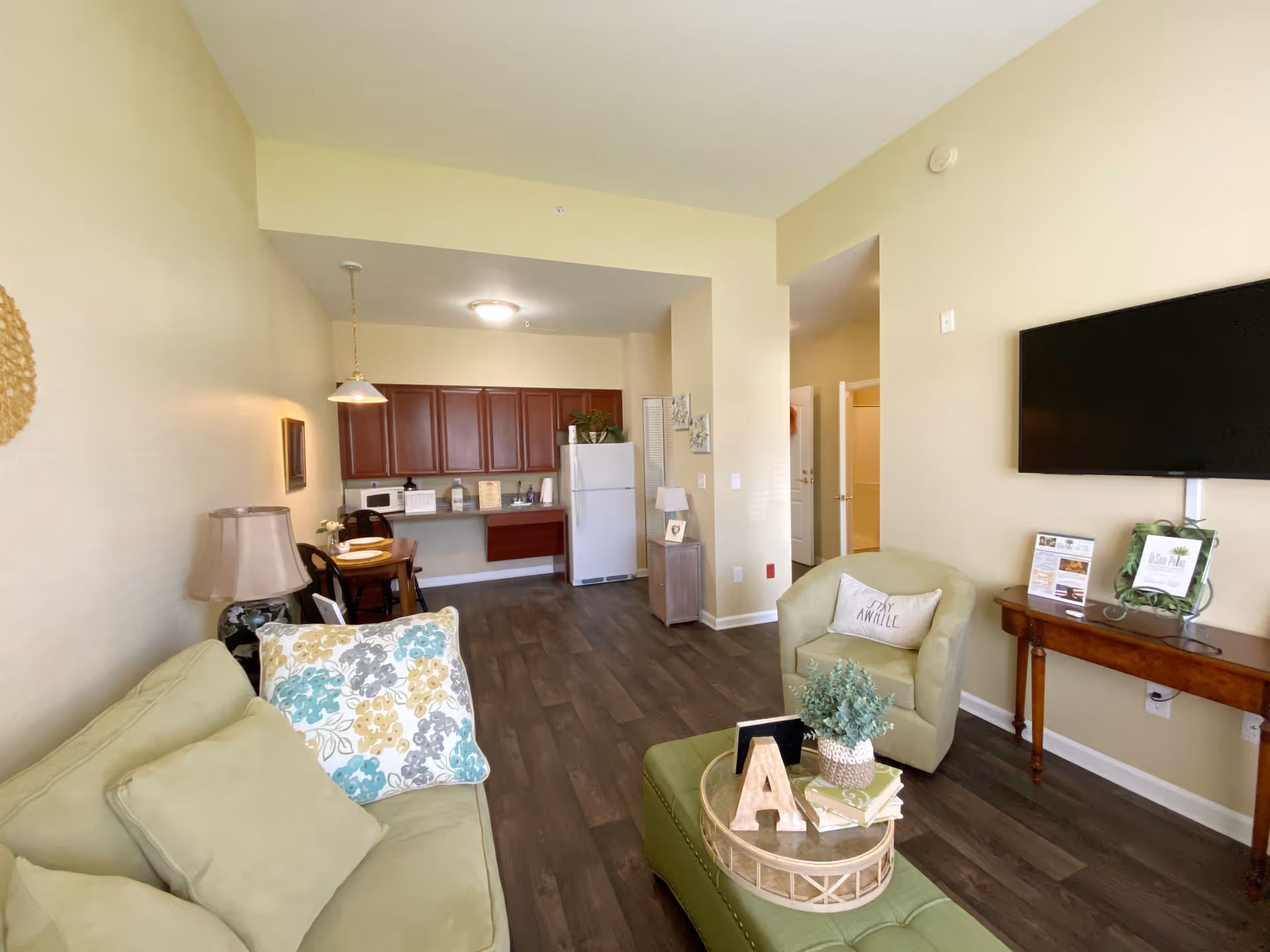 A cozy living area in an assisted living community featuring a light green sofa with patterned pillows, a matching armchair with a decorative pillow, a green ottoman with a tray holding a plant and decorative items, a wall-mounted flat-screen TV above a wooden console table with brochures, and a small dining area with a table and chairs near a kitchen with wooden cabinets and a white refrigerator.