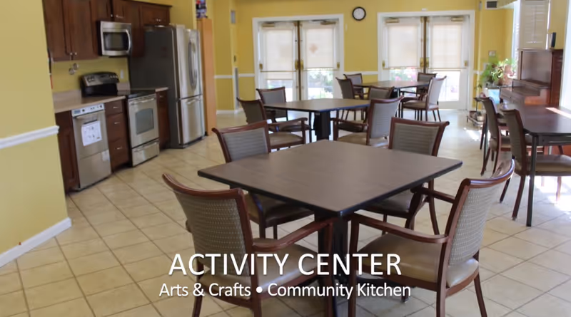 A bright activity center with several square tables and chairs arranged for group activities. The room features a community kitchen area with stainless steel appliances including a refrigerator, stove, microwave, and dishwasher. The walls are painted yellow, and there are large windows and glass doors letting in natural light. A piano is visible on the right side near the windows.