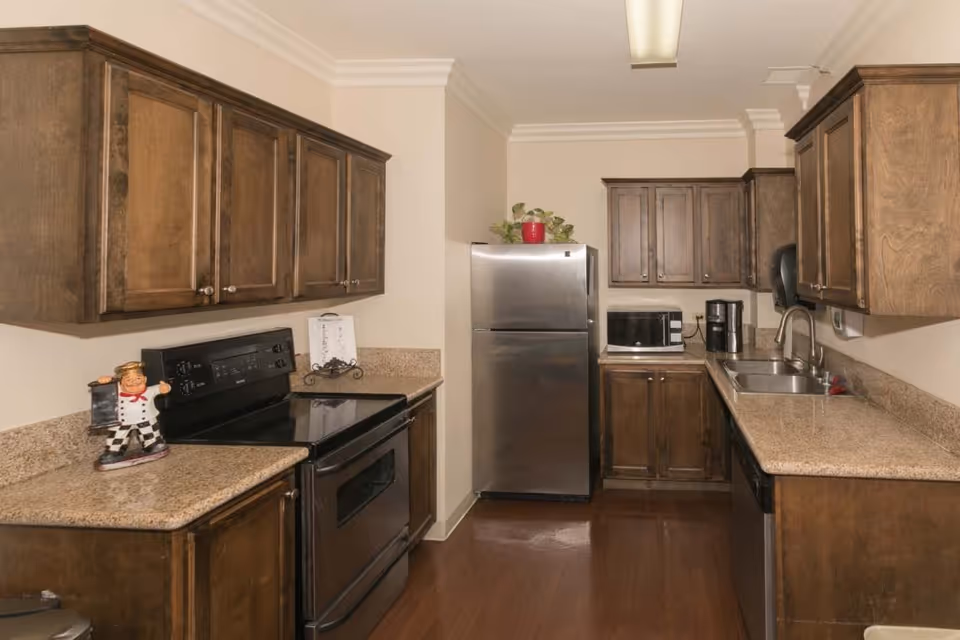 A kitchen with wooden cabinets, granite countertops, a stainless steel refrigerator, a black electric stove, a microwave, a coffee maker, and a double sink. There is a decorative chef figurine on the counter and a small plant on top of the refrigerator.