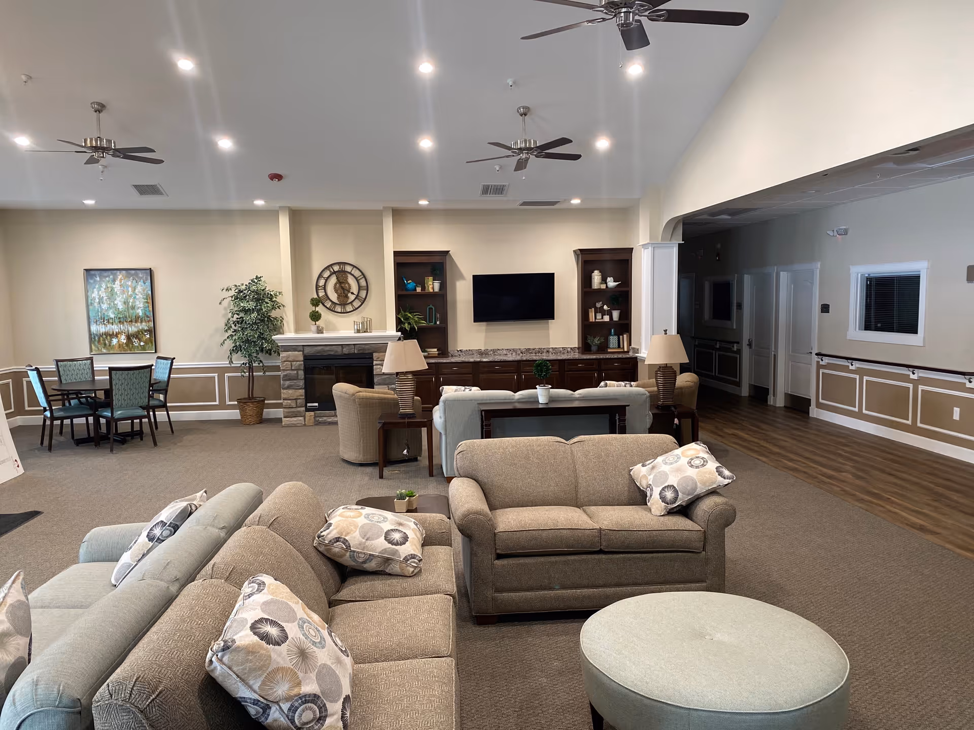 A spacious and well-lit living room area in a senior living facility featuring multiple beige sofas with patterned cushions, a round ottoman, a fireplace with a clock above it, built-in shelves, a flat-screen TV mounted on the wall, ceiling fans, and a small dining table with chairs in the background.