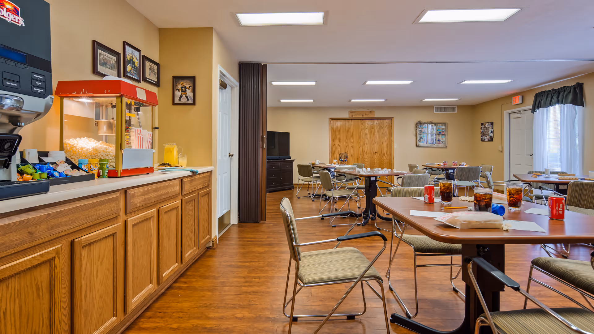 Interior view of a dining room in a senior living facility with wooden floors, several tables and chairs, a popcorn machine, and a coffee station along one wall. The room is well-lit with ceiling lights and has windows with curtains allowing natural light in.