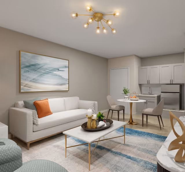 Modern open-plan living area with a white sofa, coffee table, small dining table and a kitchenette in the background.
