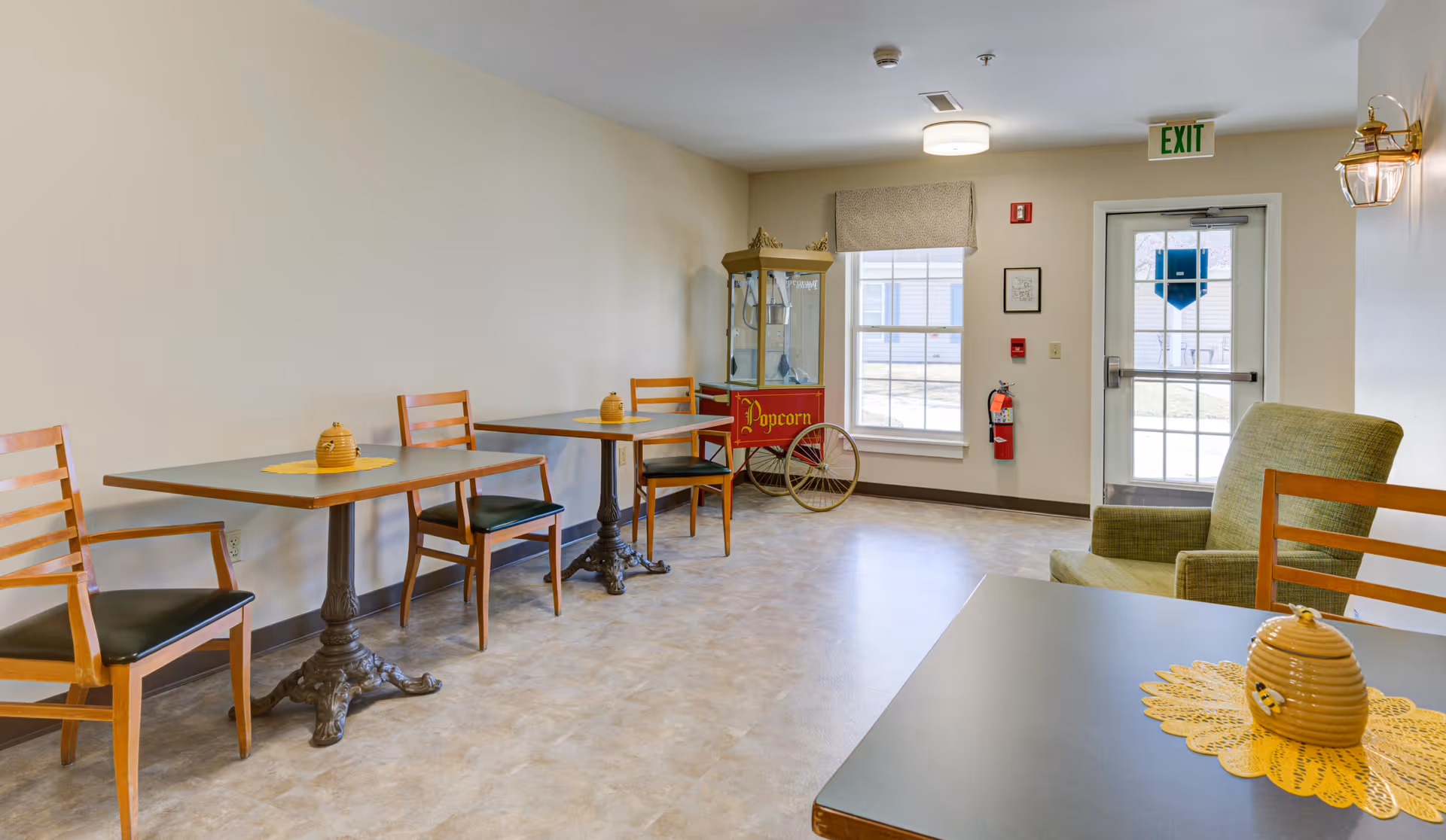 A small seating area in a senior living community with three tables, each accompanied by wooden chairs with black cushions. On each table is a yellow beehive-shaped decorative item placed on a yellow doily. In the background, there is a vintage-style popcorn machine near a window and a glass door with an exit sign above it. A green upholstered armchair is positioned near the door, and the room has light-colored walls and flooring.