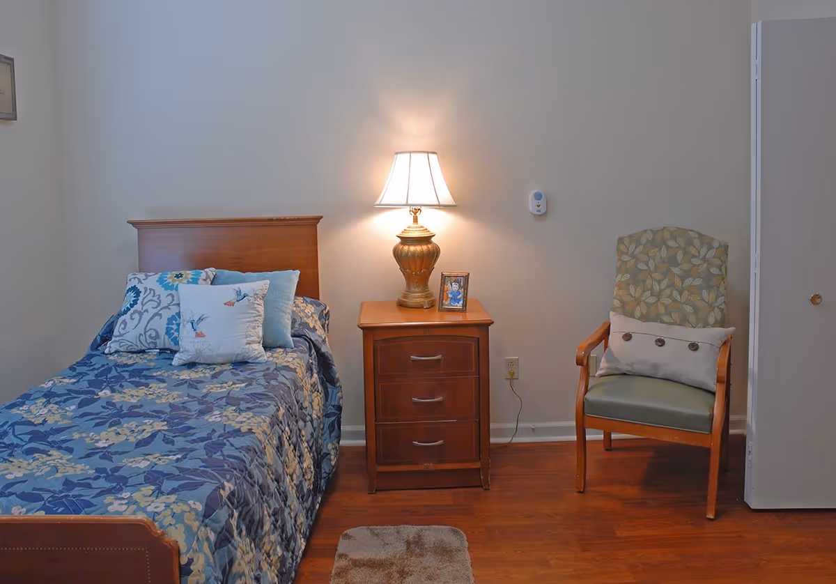 A cozy single bedroom with a bed, patterned bedding, a wooden nightstand with a lamp and photo, and an upholstered armchair.