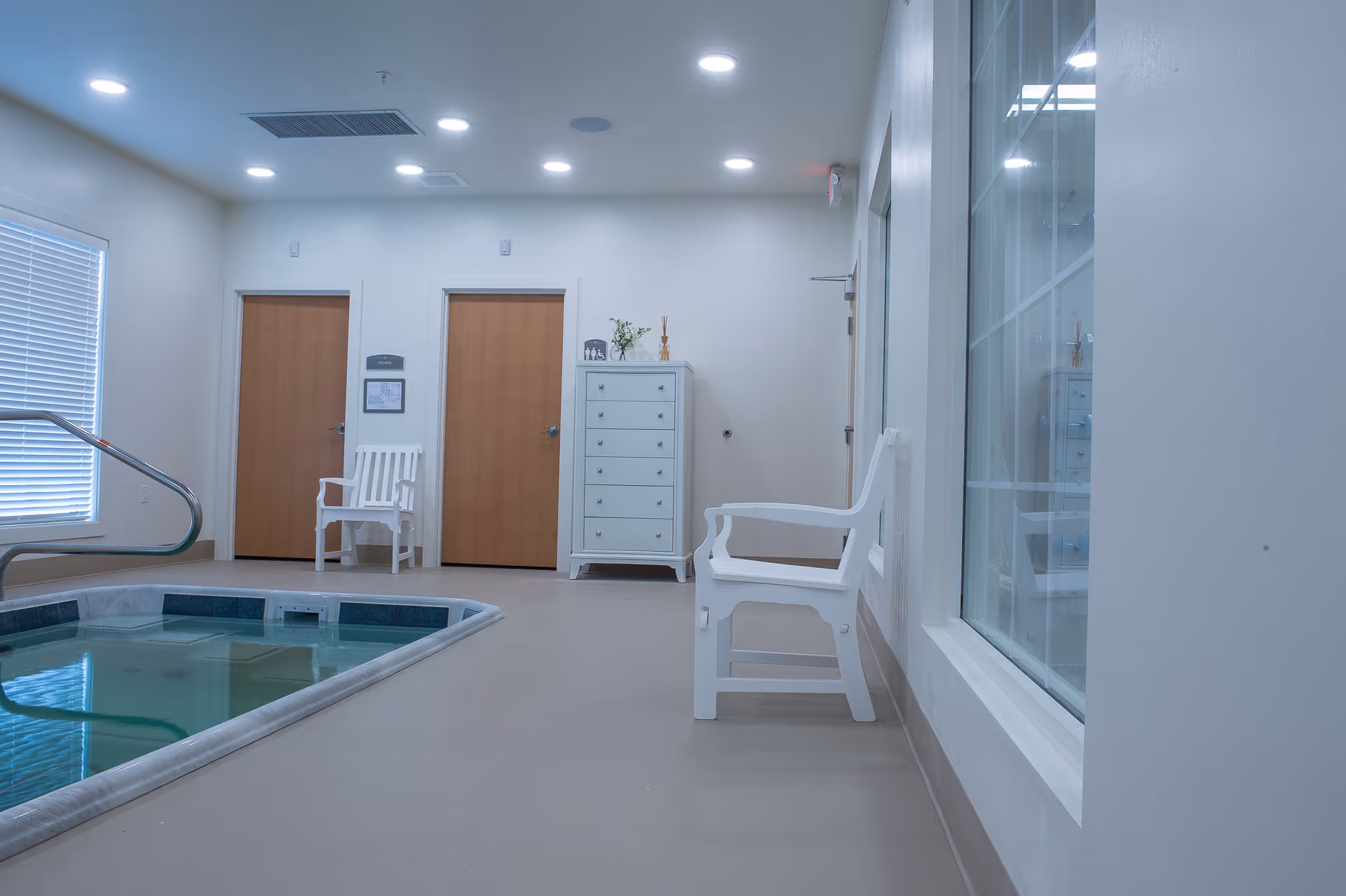 Indoor therapy pool room with a small pool, white chairs, a dresser and two wooden doors.