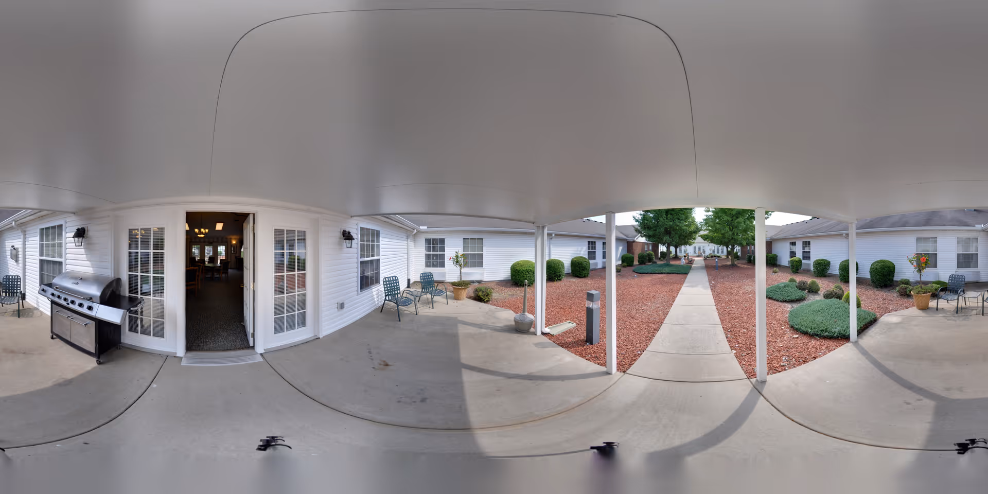 Covered outdoor patio area with a concrete floor, metal chairs, and a large barbecue grill. The patio overlooks a landscaped courtyard with red mulch, trimmed bushes, and a concrete walkway lined with trees leading to other buildings.