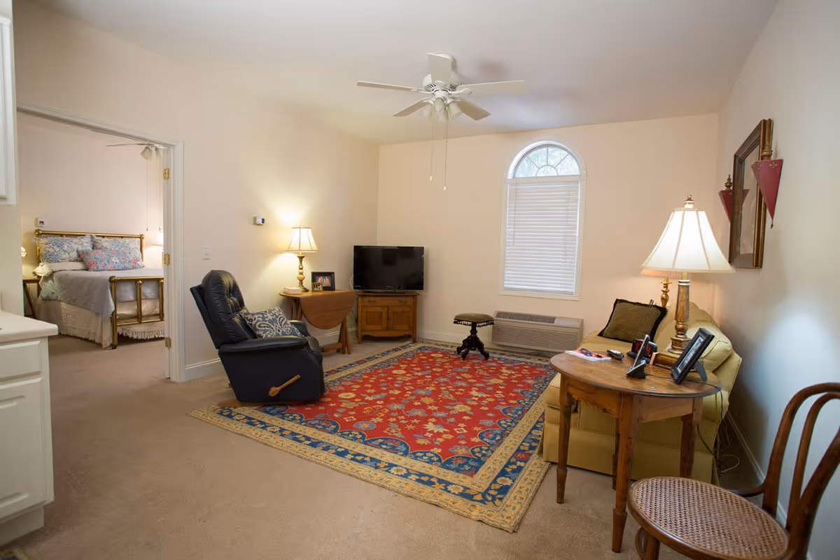 A cozy living room with a red patterned area rug, a yellow sofa with a side table and lamp, a black recliner chair, a small wooden stool, and a TV on a wooden stand. There is a window with white blinds and an arched top. A doorway leads to a bedroom with a brass bed frame and floral bedding.