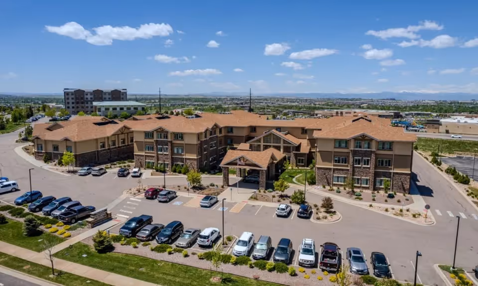 Aerial view of Pine Grove Crossing, a large senior living facility with a multi-wing building featuring a tan and brown exterior with a covered entrance. The parking lot in front is partially filled with cars, and the surrounding area includes landscaped greenery and a clear blue sky with scattered clouds.