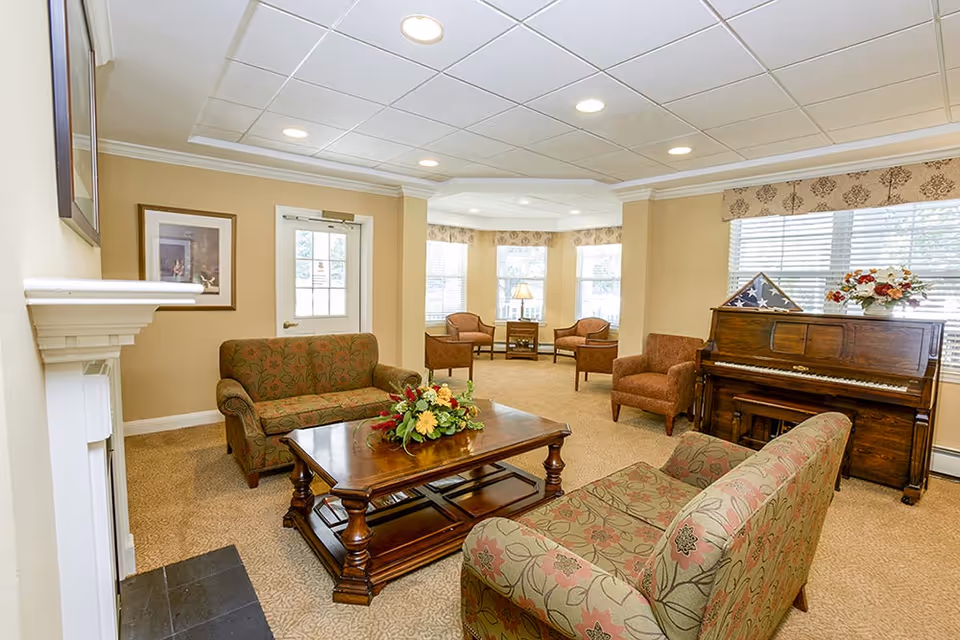A cozy living room area with two floral patterned sofas facing a wooden coffee table adorned with a flower arrangement. There is a wooden piano with a folded American flag and a flower vase on top, and several armchairs near large windows with blinds and valances. The room has beige walls, carpeted floor, recessed ceiling lights, and a white fireplace on the left side.