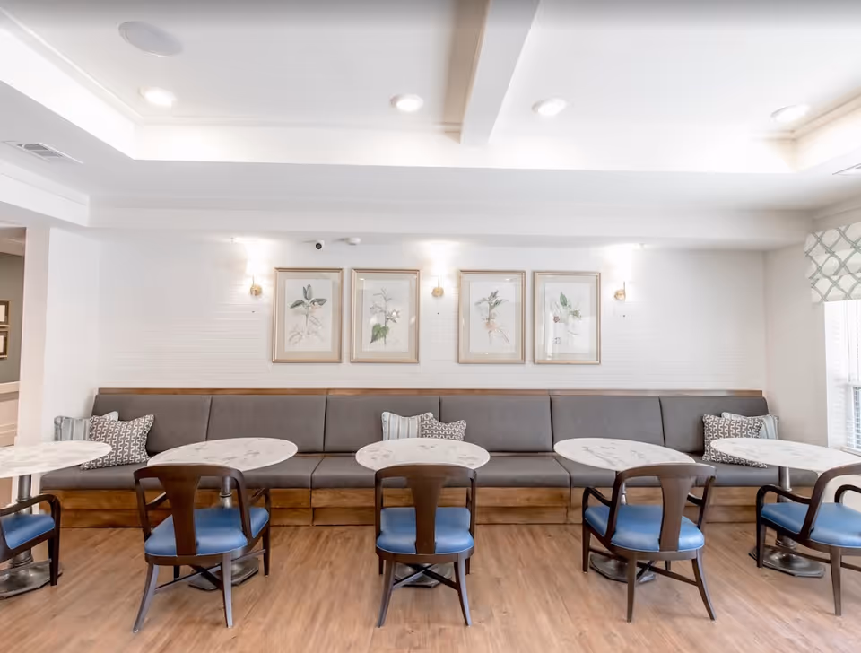 A bright dining area with four round marble tables, each paired with two wooden chairs with blue cushions. Along the back wall is a long gray cushioned bench with decorative pillows. Above the bench are four framed botanical prints, and the walls are white with soft lighting fixtures. The floor is wooden, and there is a window with patterned curtains on the right side.