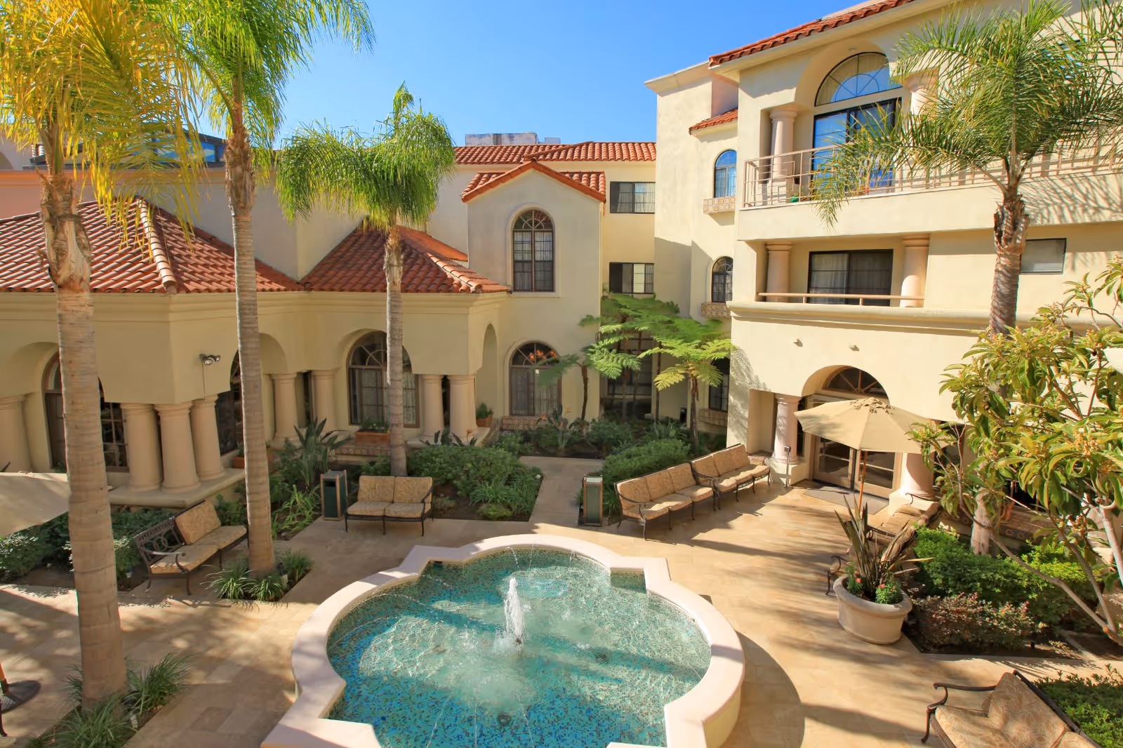 A sunny courtyard at Palm Court featuring a central decorative fountain surrounded by benches and lush greenery, with palm trees and Mediterranean-style buildings with red tile roofs and arched windows in the background.