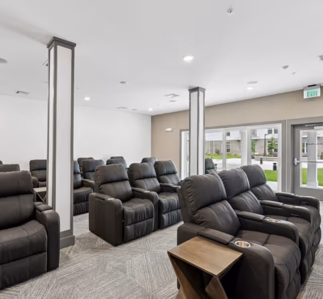 A modern lounge area with multiple rows of black leather recliner chairs arranged facing forward. The room has light-colored walls, carpeted flooring, and large windows letting in natural light. There are two white pillars with gray trim in the middle of the room and a small wooden side table with a built-in cup holder next to one of the recliners.