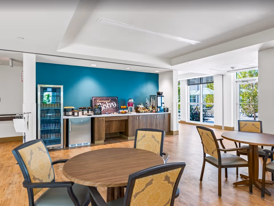 Bright communal dining area with round tables and chairs facing a beverage and snack station against a teal accent wall and large windows.