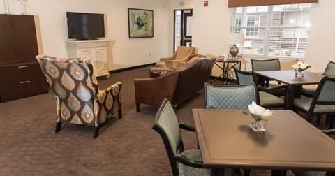 A cozy common area with patterned armchairs, a brown leather sofa, and several tables with green cushioned chairs. There is a flat-screen TV mounted on a white fireplace mantel, a painting on the wall, and a window with blinds letting in natural light.