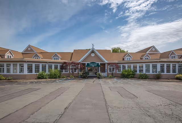 Single-story tan senior living building with a peaked central entrance, rows of windows, shrubs, and an empty asphalt parking lot under a partly cloudy sky.