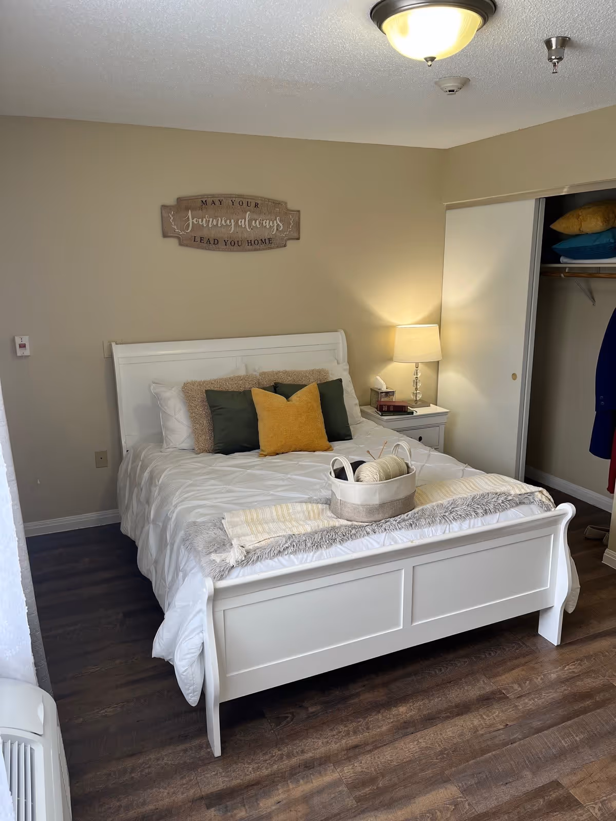 A cozy bedroom with a white wooden bed frame, white bedding, and multiple pillows in green, yellow, and beige colors. A small white nightstand with a lamp, books, and a tissue box is beside the bed. Above the bed is a wooden wall sign that reads 'May your journey always lead you home.' There is an open closet with sliding doors showing some folded blankets and hanging clothes. The floor is wooden, and the room has beige walls.