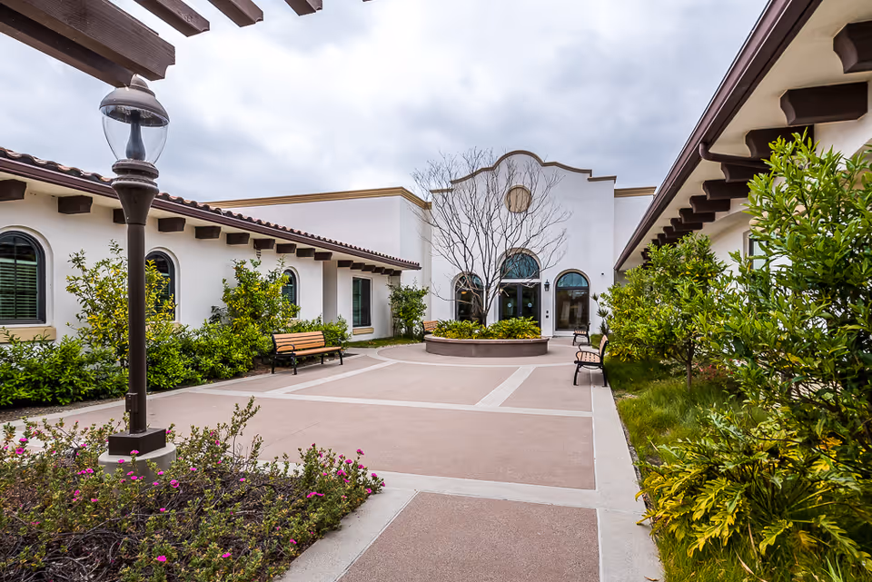 Outdoor courtyard area at Oceanside Senior Living with benches, a central planter with a leafless tree, surrounding greenery, and white buildings with arched windows under a cloudy sky.
