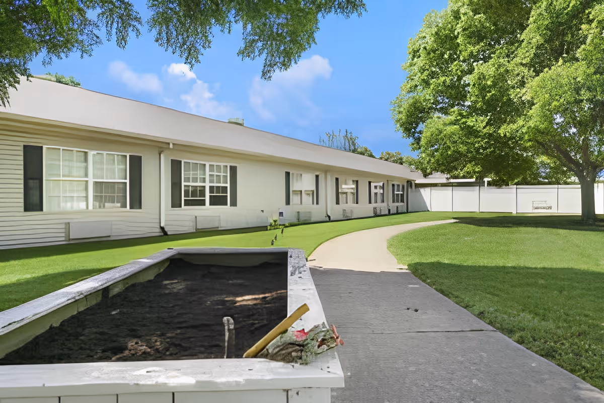Outdoor view of a senior living facility with a paved walkway curving through a well-maintained grassy area. The building has white siding with multiple windows and black shutters. Large green trees provide shade, and a white fence encloses the area in the background.