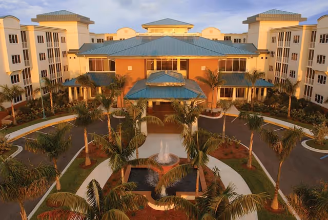 Front entrance of a multi-story senior living building with blue-green metal roofs, a circular driveway lined with palm trees and a central fountain.
