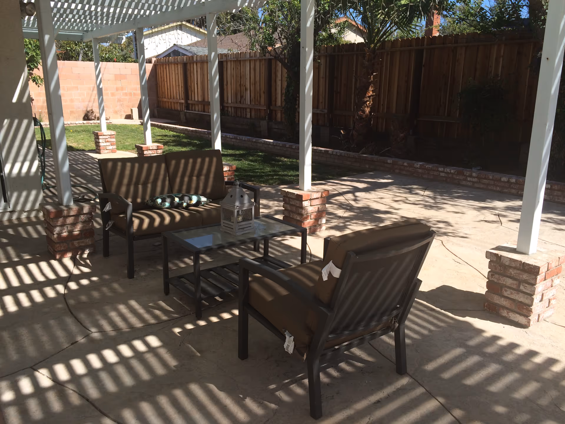 Outdoor patio area with a pergola casting striped shadows on the concrete floor. The patio has a seating arrangement with a cushioned loveseat, a cushioned chair, and a glass-top coffee table with a decorative lantern and a bowl of ornaments. The area is enclosed by a wooden fence with some greenery and trees visible.