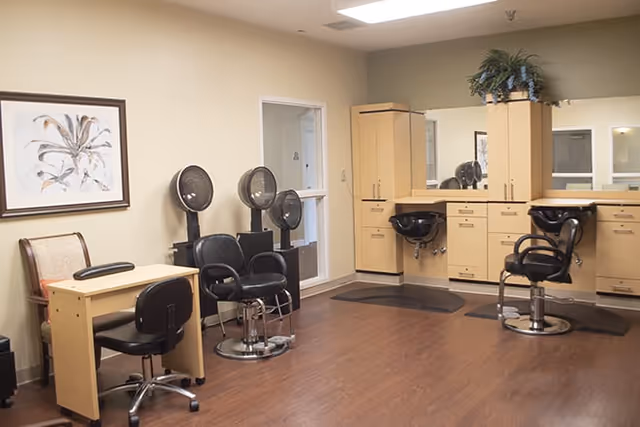 Interior of a hair salon area with wooden flooring, beige walls, two black salon chairs in front of a counter with sinks and mirrors, two hair dryers on stands, a small desk with a chair, and a framed botanical artwork on the wall.