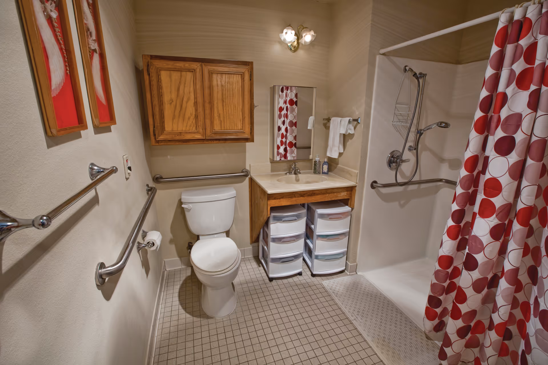 A bathroom with a toilet, a sink with a mirror above it, and a shower area with a red and white polka dot shower curtain. The bathroom has beige walls, a tiled floor, wooden cabinets above the toilet, and grab bars installed near the toilet and in the shower for accessibility.