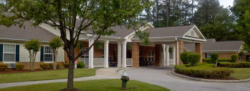Front exterior view of a single-story senior living facility building with a covered entrance supported by brick columns, surrounded by green trees, shrubs, and a well-maintained lawn.
