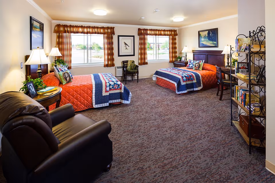 A cozy bedroom with two twin beds covered in orange quilts and blue patterned blankets. Each bed has a pillow with a dog image. The room features two windows with plaid curtains, a leather armchair, a round side table with a lamp and framed photo, a wooden desk with a lamp, and a metal bookshelf filled with books. The walls are decorated with framed artwork.
