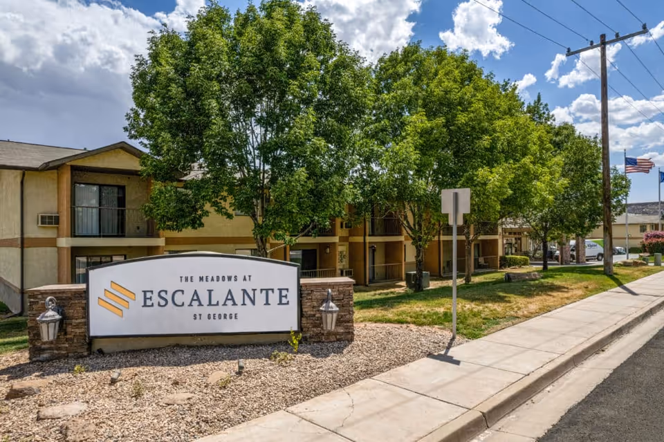 Exterior view of The Meadows at Escalante facility with a large sign in the foreground displaying the facility name. The building is two stories with balconies and surrounded by green trees and a sidewalk along the street. The sky is partly cloudy.