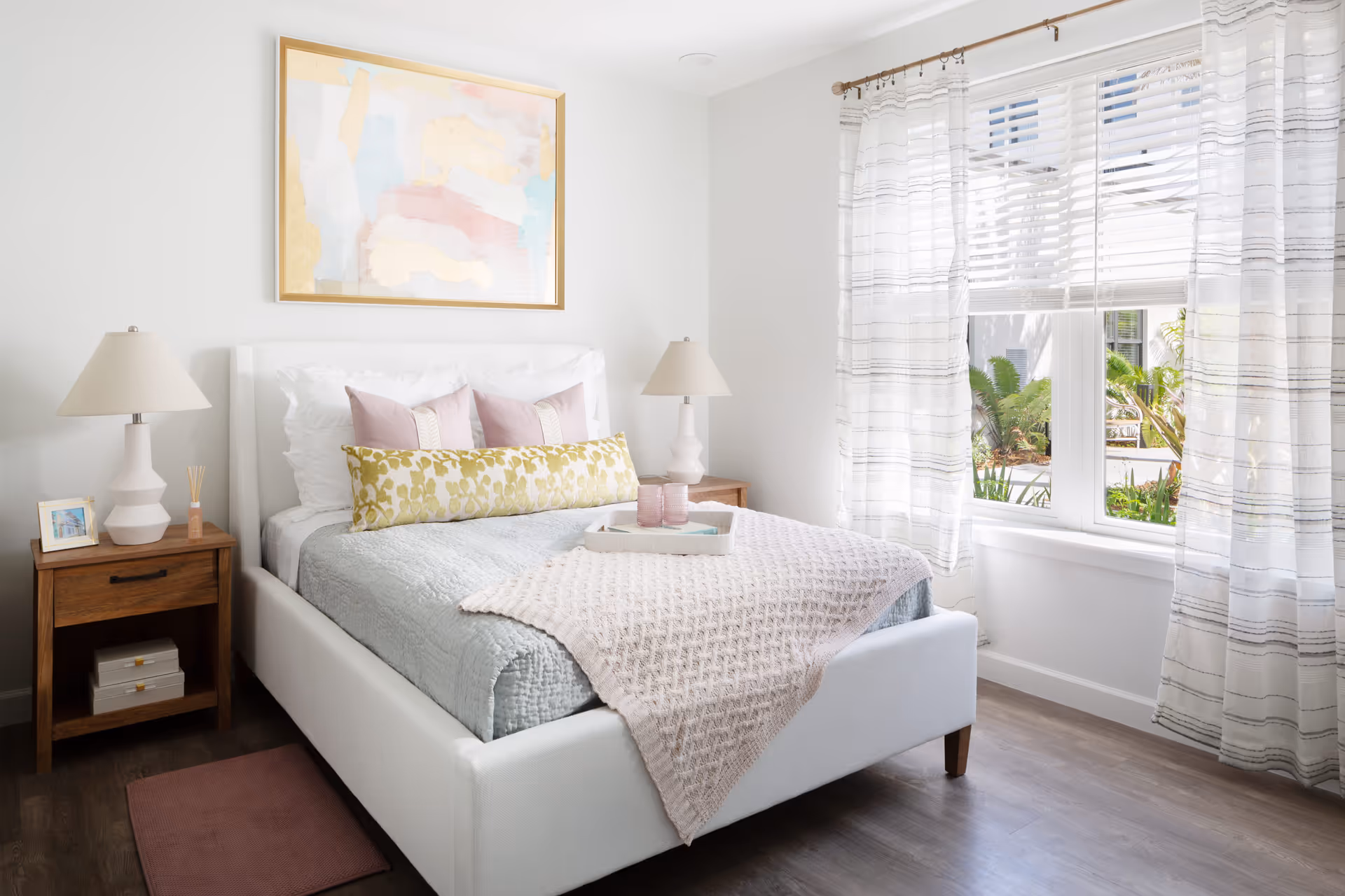Bright, neatly made bedroom with a white upholstered bed, pastel pillows and throw, wooden nightstands with lamps, and a large window with sheer curtains.