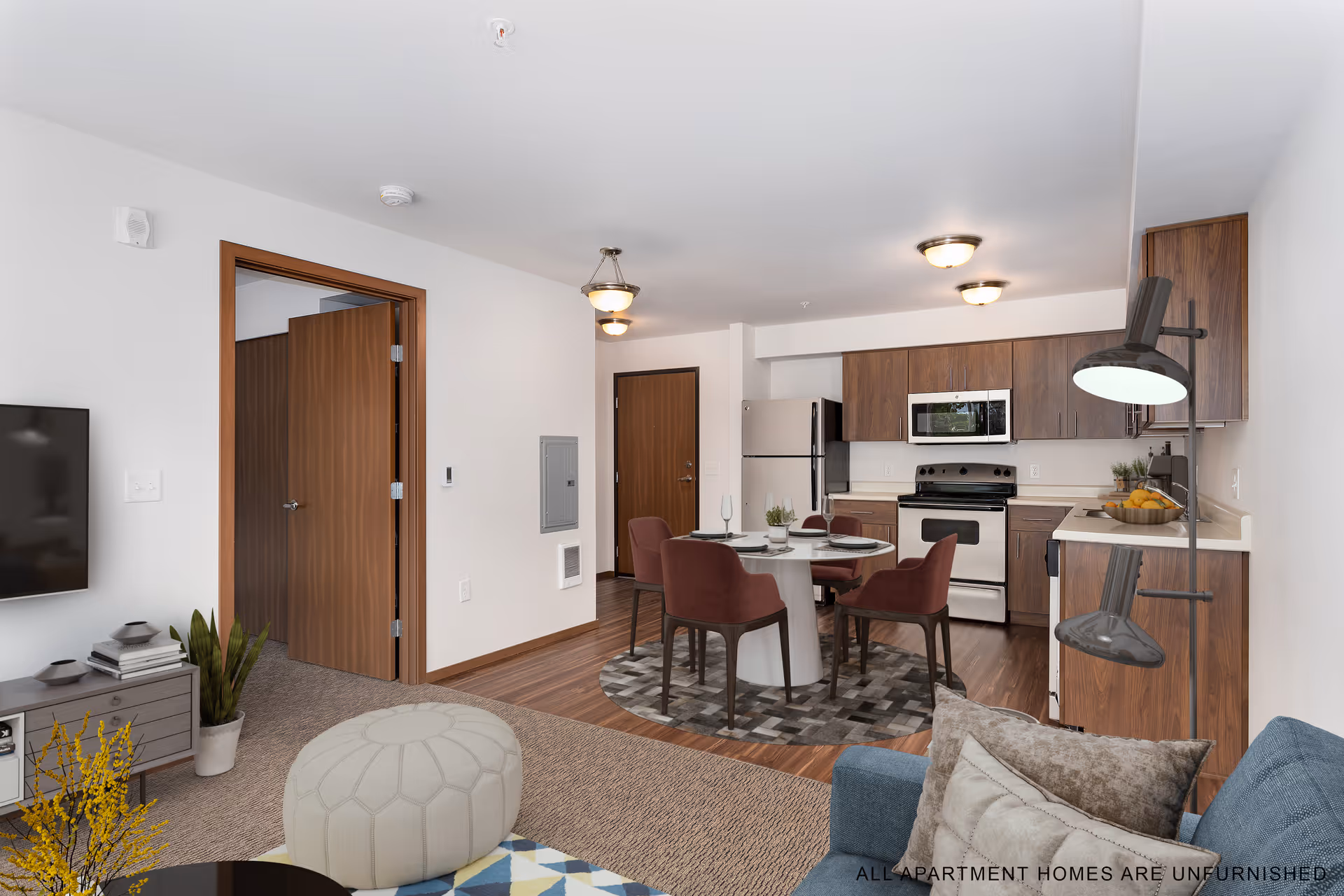 A modern open-concept living space featuring a kitchen with wooden cabinets, stainless steel appliances including a refrigerator, stove, and microwave. In front of the kitchen is a round dining table with four burgundy chairs on a patterned rug. The living area includes a blue sofa with pillows, a beige pouf, a small table with books and plants, and a wall-mounted TV. The room has wood flooring in the kitchen and carpet in the living area. There are two wooden doors visible, one open and one closed. The walls are white, and ceiling lights illuminate the space.