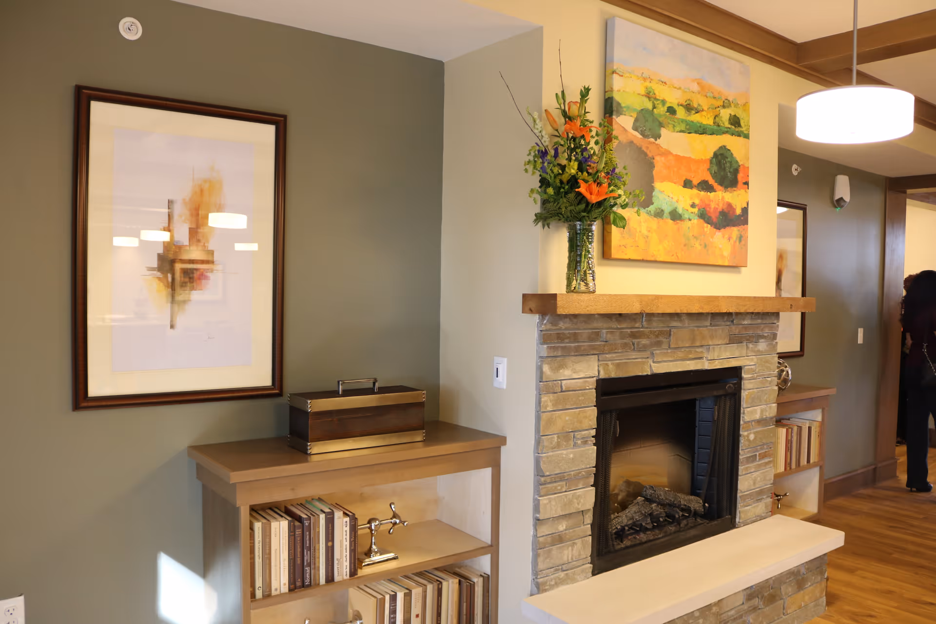 A cozy interior corner of a senior living facility featuring a stone fireplace with a wooden mantel. On the mantel is a vase with a colorful floral arrangement. Above the mantel hangs a bright landscape painting. To the left of the fireplace is a wooden bookshelf with books and decorative items, and a framed abstract artwork hangs on the wall above it. The room has warm lighting and wooden flooring.
