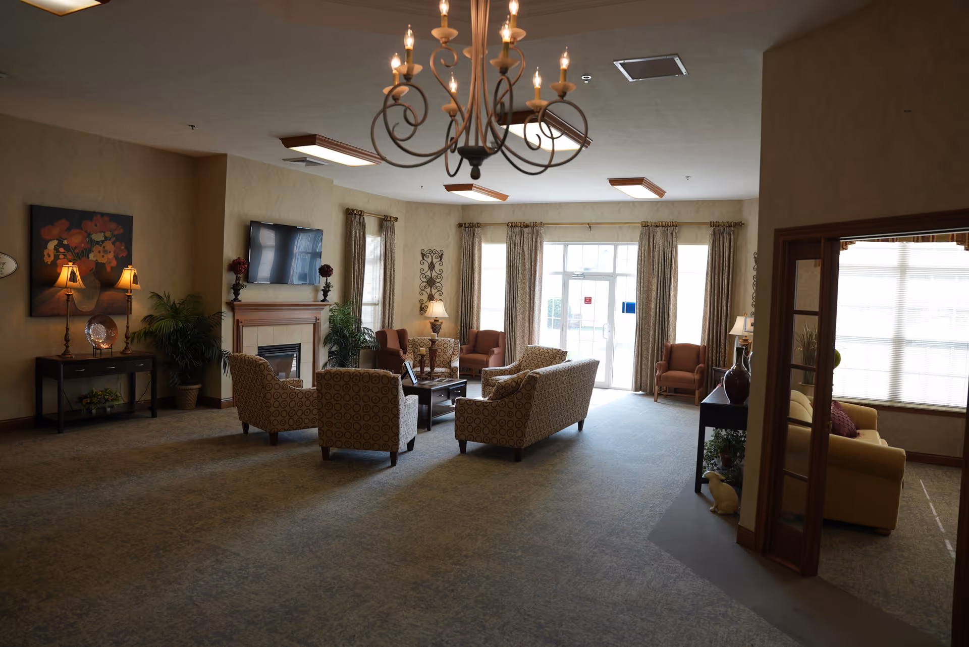 A spacious and well-lit living room area in Brownsburg Meadows featuring multiple patterned armchairs and sofas arranged around a central coffee table. The room includes a fireplace with a mounted flat-screen TV above it, decorative lamps, plants, and large windows with curtains allowing natural light to enter. A chandelier hangs from the ceiling, and there is an adjacent smaller seating area visible through a doorway on the right.