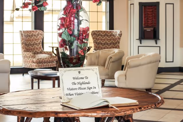 A lobby-style seating area with upholstered chairs around a wooden table holding a guest sign-in book and a tall vase of red flowers.