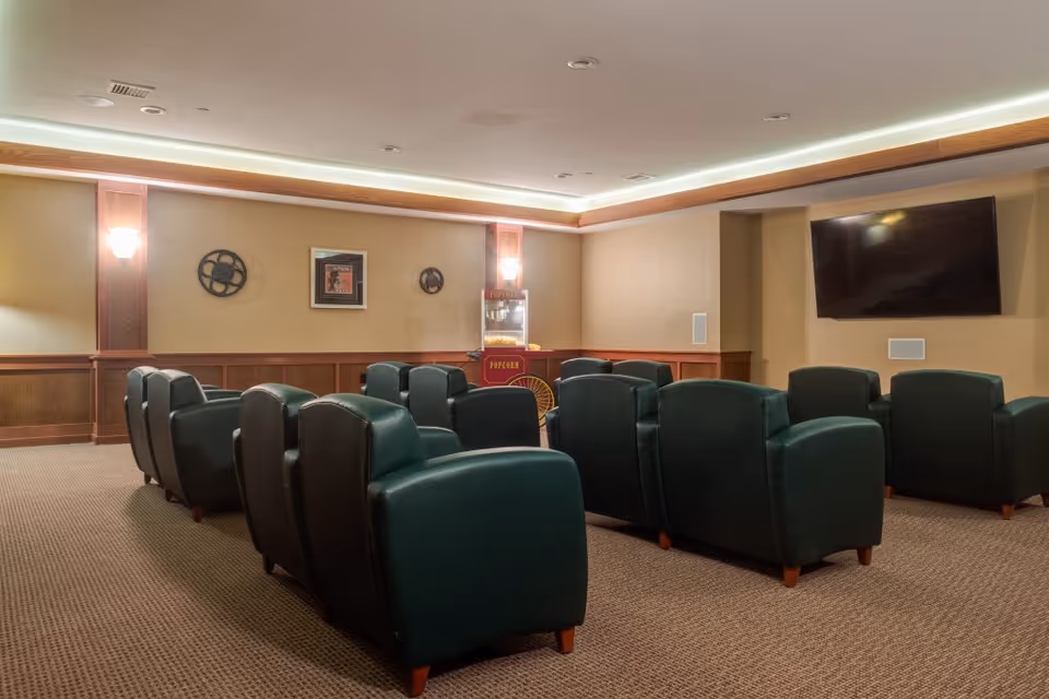 A cozy senior living facility media room with two rows of dark green leather armchairs facing a wall-mounted flat screen TV. The room features warm beige walls with wooden paneling, soft wall sconces, movie-themed decorations, and a vintage-style popcorn machine in the corner.