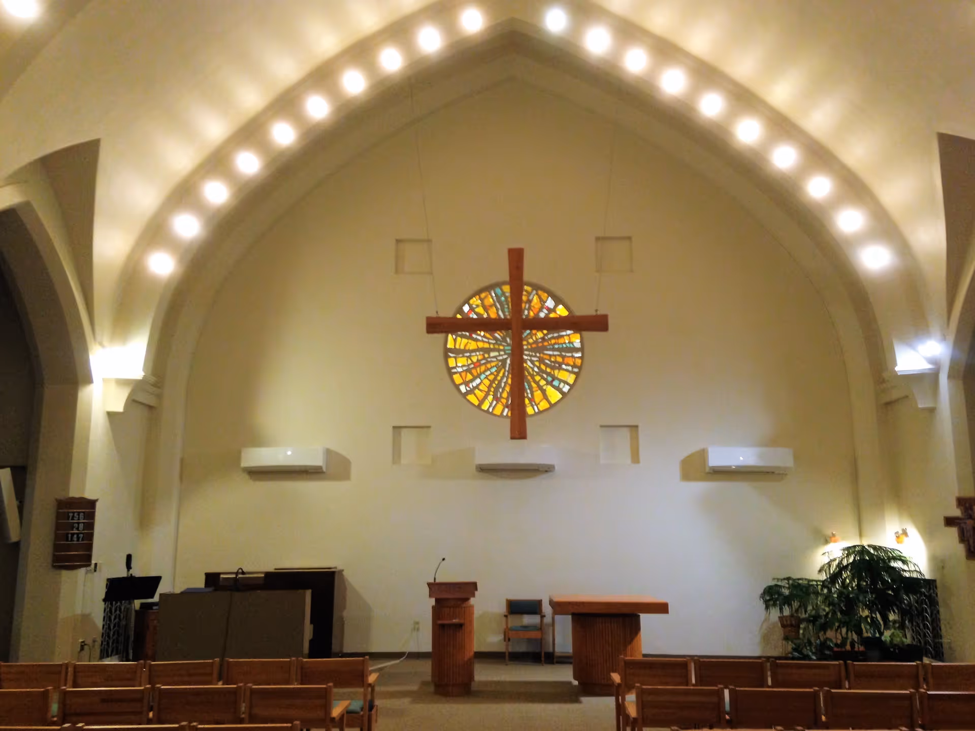 Interior view of a chapel with a large wooden cross hanging in front of a circular stained glass window. The chapel has arched ceilings with lights along the arch, wooden pews, a lectern with a microphone, a table, and some plants on the right side.