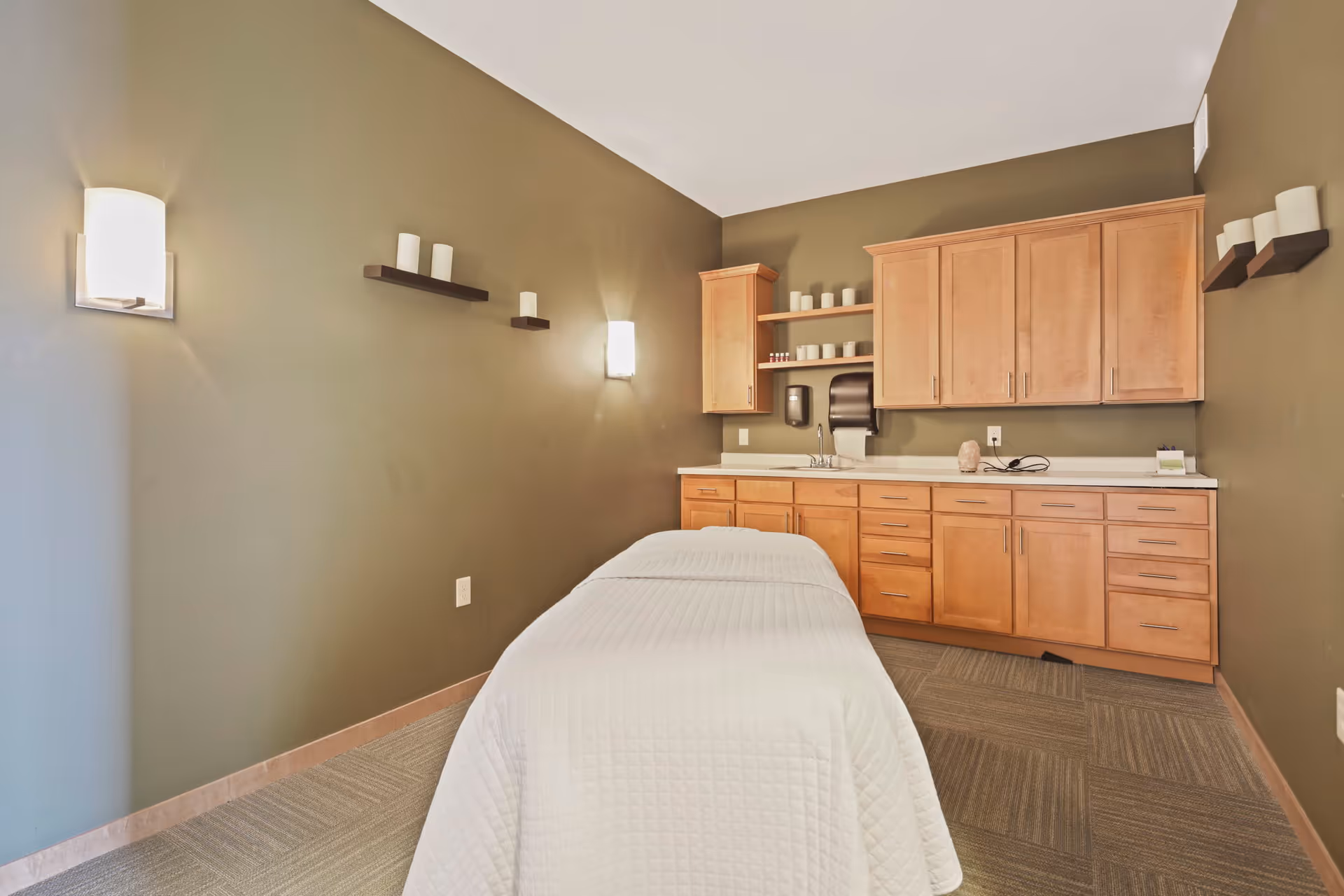 A serene treatment room with a massage table covered in a white quilted sheet, olive green walls, wooden cabinets and shelves holding candles and small containers, soft wall-mounted lights, and a carpeted floor.
