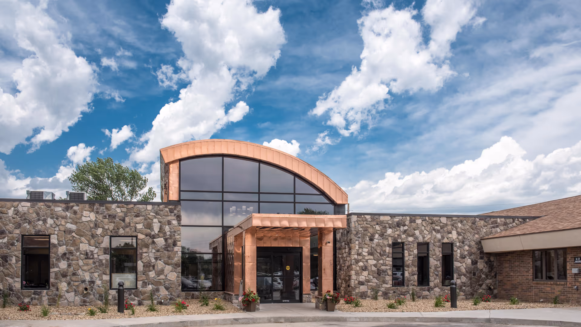 Front exterior view of a healthcare facility building with stone and brick walls, large arched glass windows, and a copper-colored entrance canopy under a partly cloudy sky.