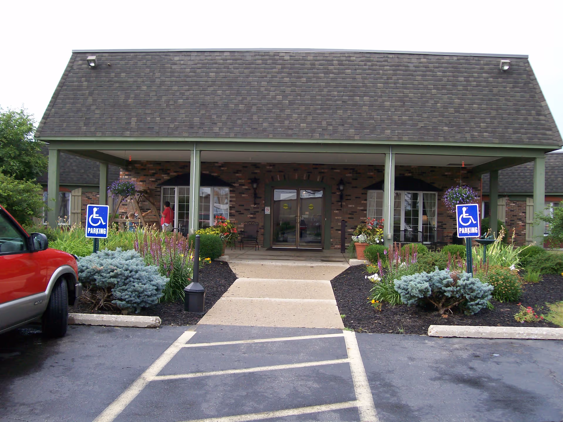 Front entrance of a brick building with a covered porch, two handicap parking signs on either side of the walkway, landscaped flower beds, and a red vehicle parked on the left side.