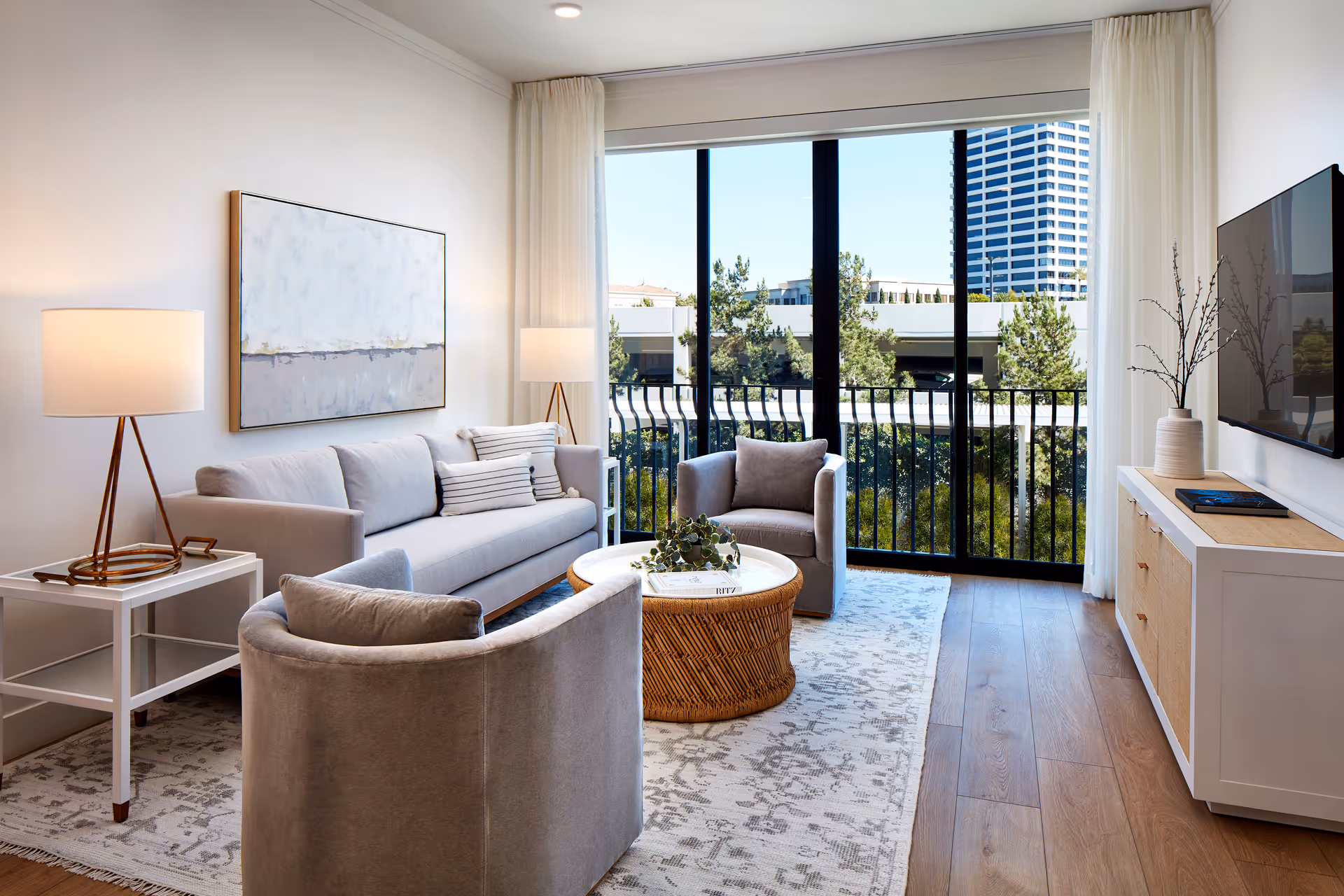 Modern bright living room with a sofa, two armchairs, a round wicker coffee table, and floor-to-ceiling windows overlooking a balcony and city view.