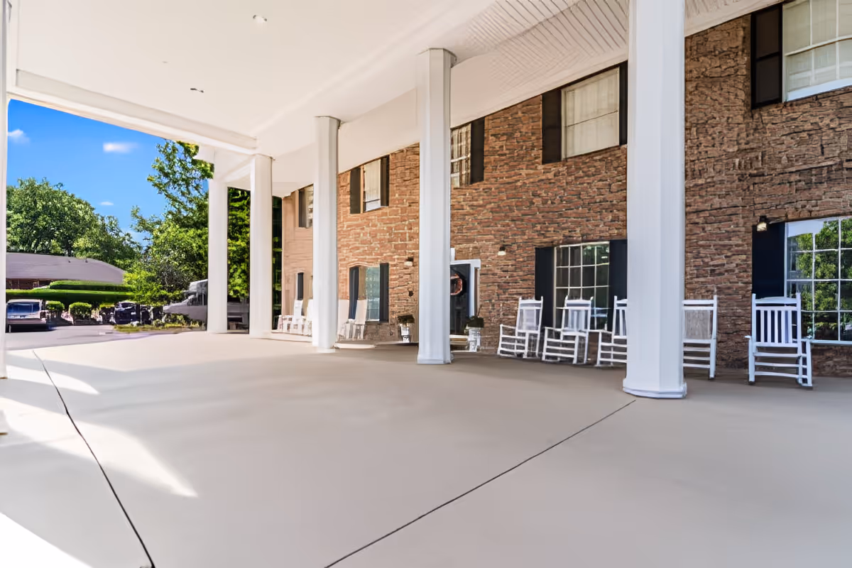 Covered outdoor entrance area of a building with large white columns and several white rocking chairs lined up against a brick wall under the roof. Trees and parked cars are visible in the background under a blue sky.