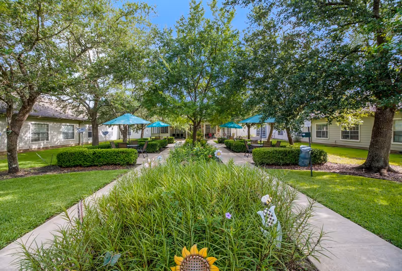 Outdoor garden area at Sodalis Texas City with a central flower bed surrounded by paved walkways, green grass, trees, and patio tables with blue umbrellas. Residential buildings are visible in the background.