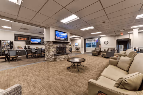 A spacious senior living common area featuring a stone fireplace with a mounted flat-screen TV above it. The room has comfortable seating including sofas and armchairs arranged around a round coffee table. In the background, there is a dining area with tables and chairs, a sign that reads 'Bistro', and another TV mounted on the wall. The room has a carpeted floor, a drop ceiling with fluorescent lighting, and an American flag near a window with blinds.