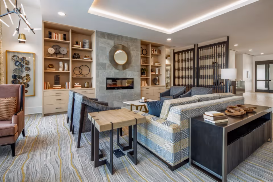 A modern living room area in a senior living facility featuring a patterned sofa, several armchairs, a wooden side table, and built-in shelves with decorative items and books. A stone fireplace with a round mirror above it is centered between the shelves. The room has a patterned carpet and soft lighting, with a decorative screen partition in the background.