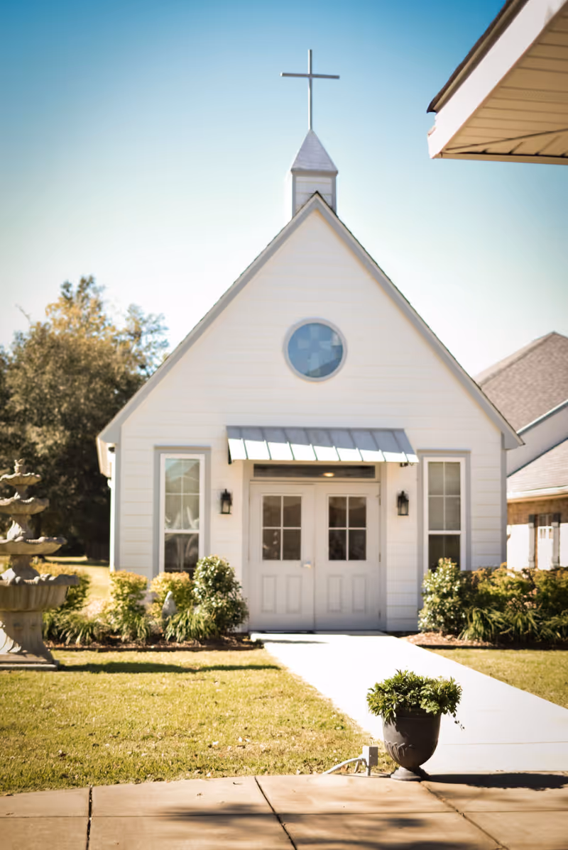 Small white chapel building with a cross on top, double doors at the entrance, flanked by two windows, surrounded by green shrubs and a lawn with a stone fountain on the left side.