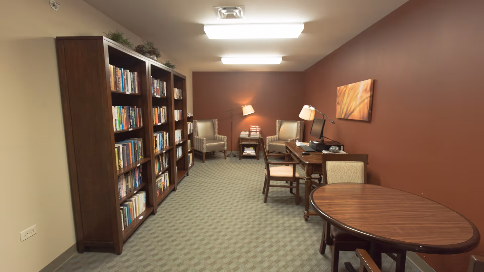 A quiet reading and work area with bookshelves filled with books on the left side, two armchairs with floor lamps in the back, a desk with a computer and chair on the right, and a round wooden table with chairs in the foreground. The walls are painted beige and dark red, and the floor is carpeted.