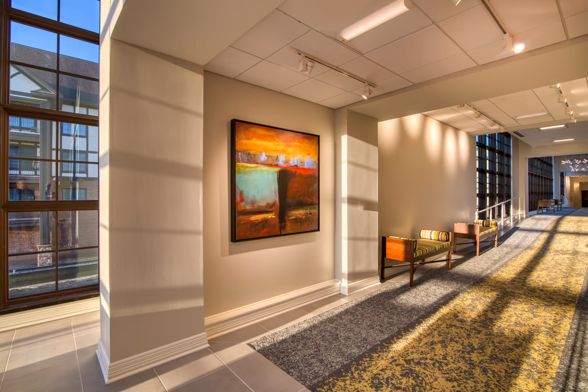 A brightly lit hallway with large windows on the left side allowing natural light to cast shadows on the walls and floor. The hallway features a colorful abstract painting on the wall and two wooden benches with striped cushions. The floor is covered with a patterned carpet, and the ceiling has recessed lighting.