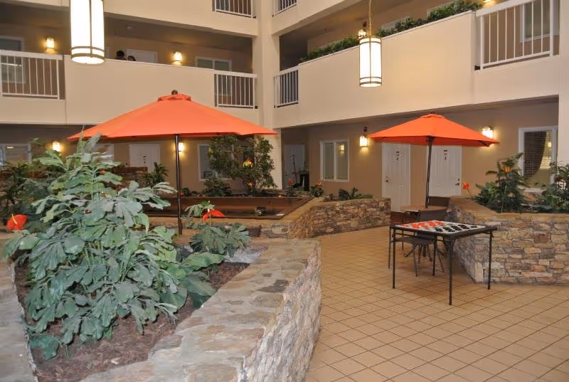 Indoor courtyard area with stone planter walls containing green plants and red flowers. Two orange umbrellas provide shade over seating areas, including a table with a checkerboard game. The space is surrounded by multiple floors with white railings and doors leading to rooms.
