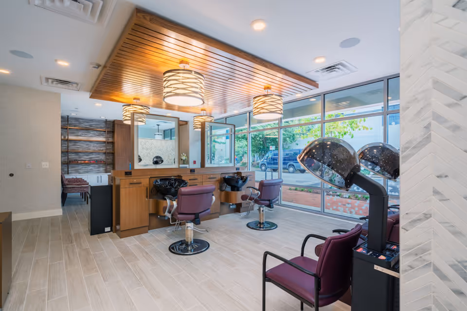 Interior view of a modern hair salon area in a senior living facility with large windows allowing natural light. The room features wooden cabinetry with mirrors, three purple salon chairs in front of hair washing sinks, and two purple chairs under hair dryers. The ceiling has wooden paneling with three hanging light fixtures.